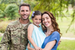 A military veteran wearing camouflage fatigues standing next to their spouse and child.