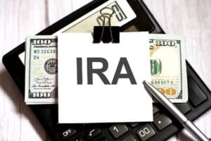 On top of a calculator, a note that reads “IRA” is paperclipped to a stack of $100 bills next to a silver pen.
